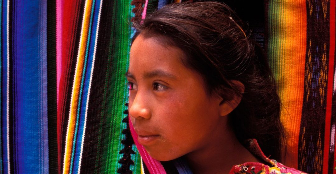 A girl peaks out from behind a colorful hanging blanket in Chichicastenango, Guatemala