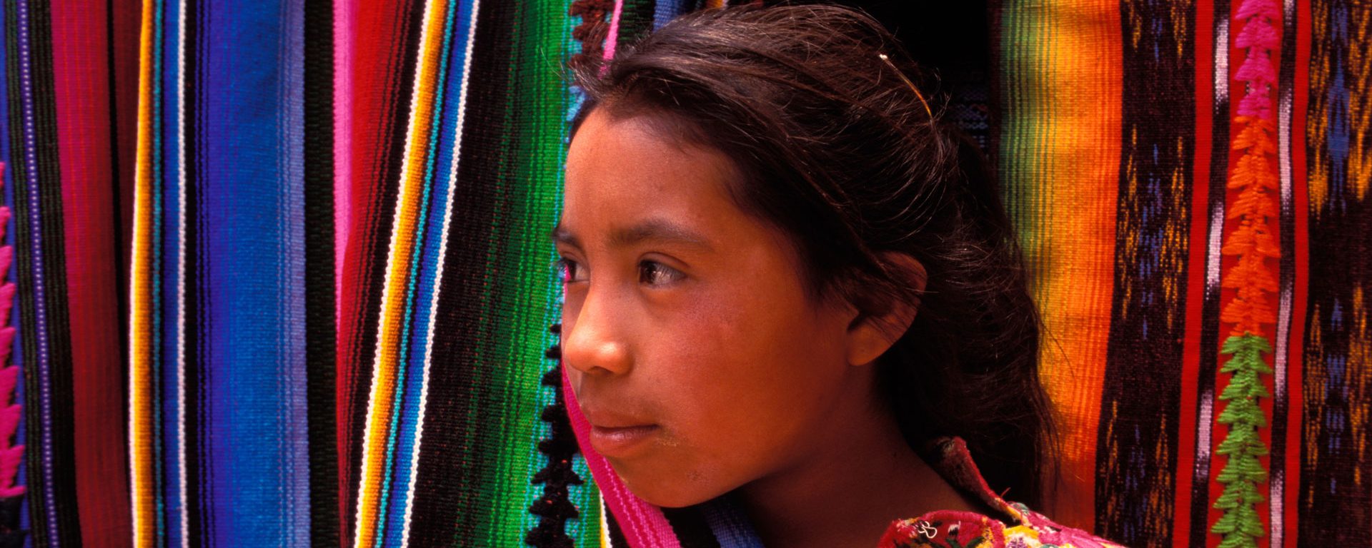 A girl peaks out from behind a colorful hanging blanket in Chichicastenango, Guatemala