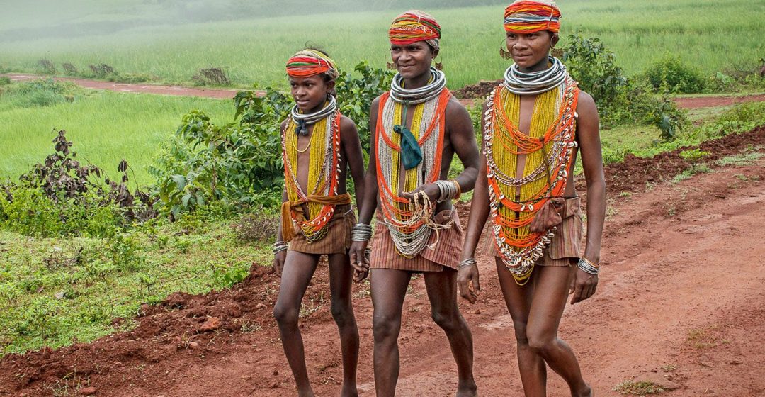 Three young Bonda women in beaded tribal dress walk down a red dirt road, India