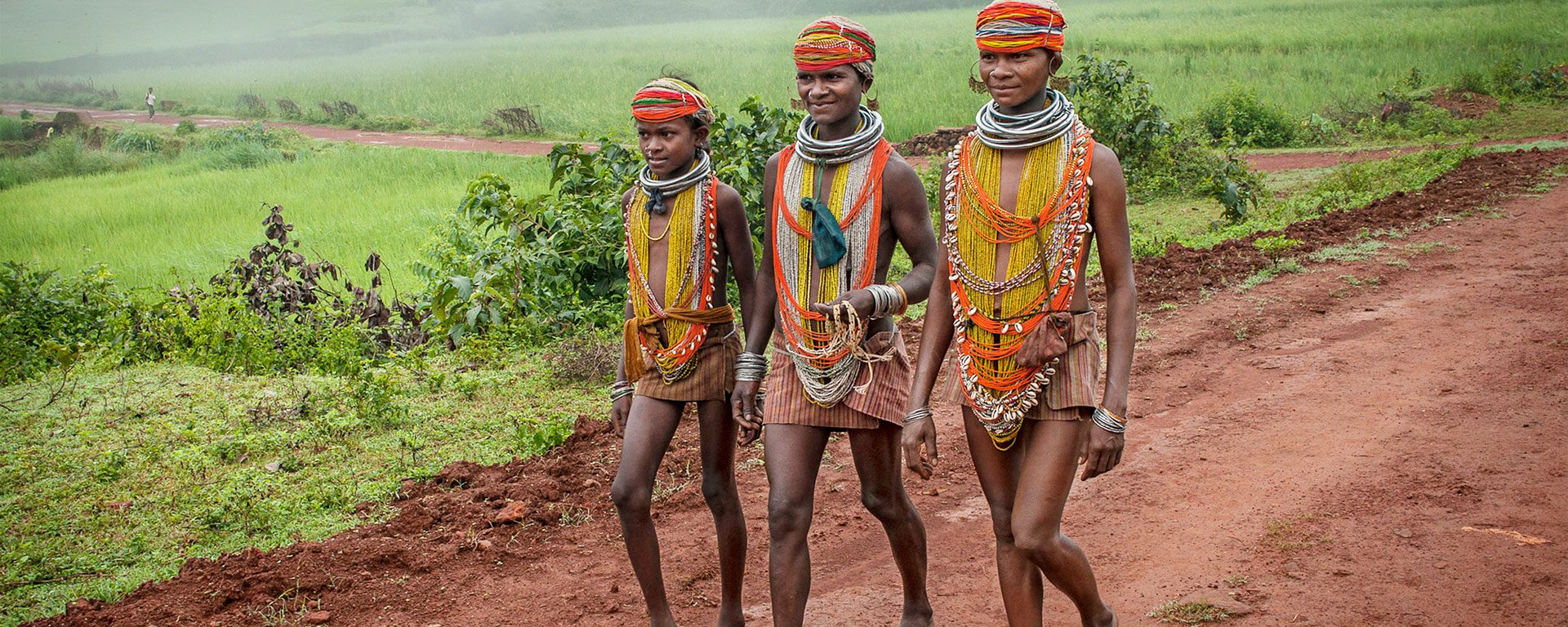 Three young Bonda women in beaded tribal dress walk down a red dirt road, India