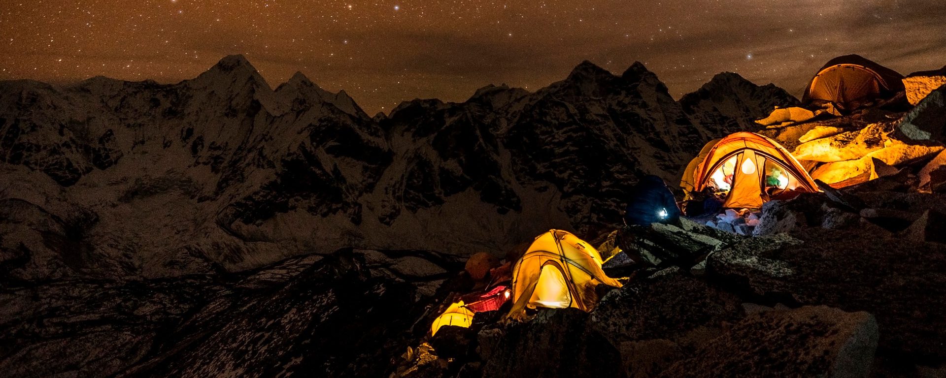 Trekking tents glowing against the night sky with mountains in the distance